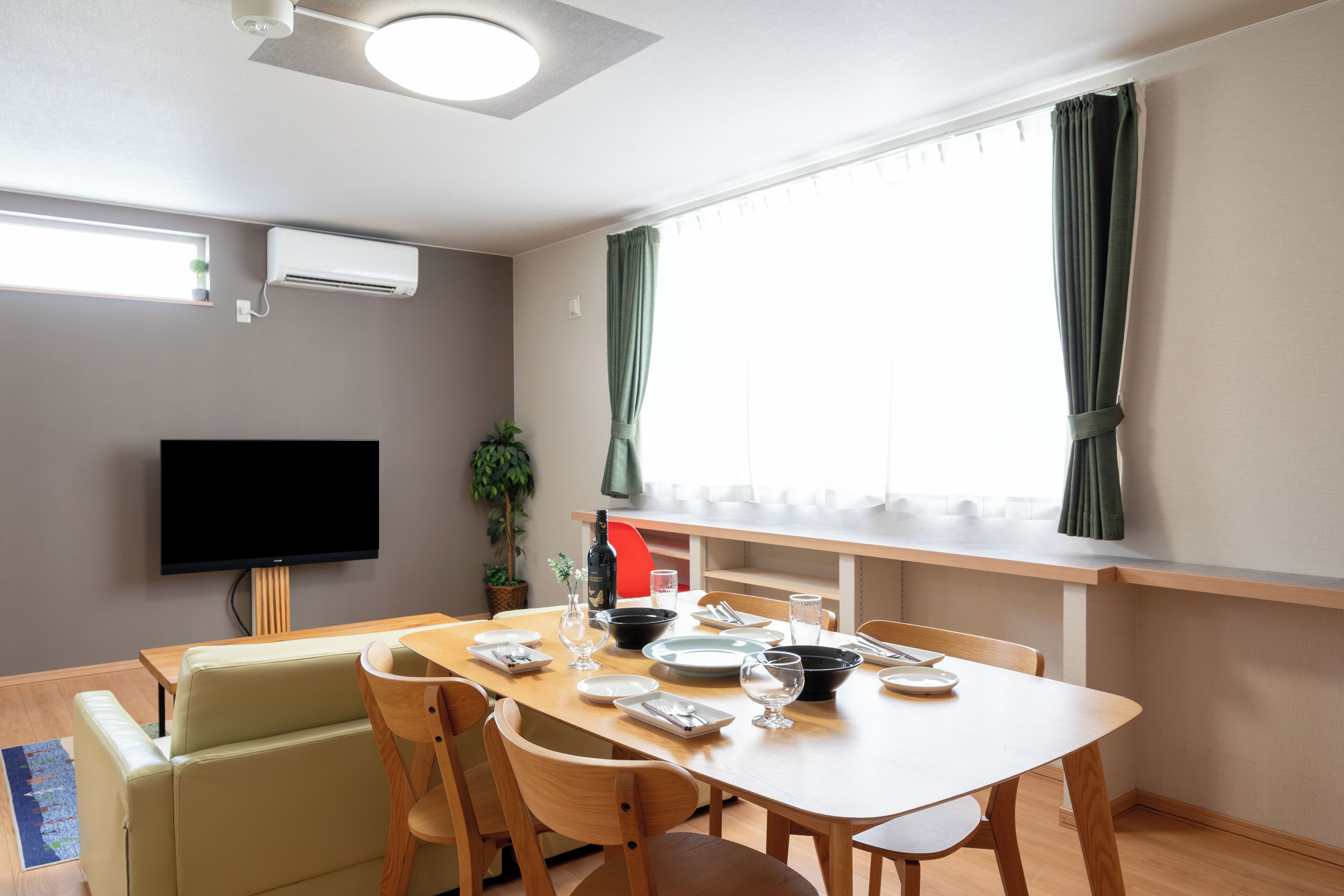 A modern dining area with a wooden table set for four, a flat-screen TV mounted on the wall, and a window with curtains.