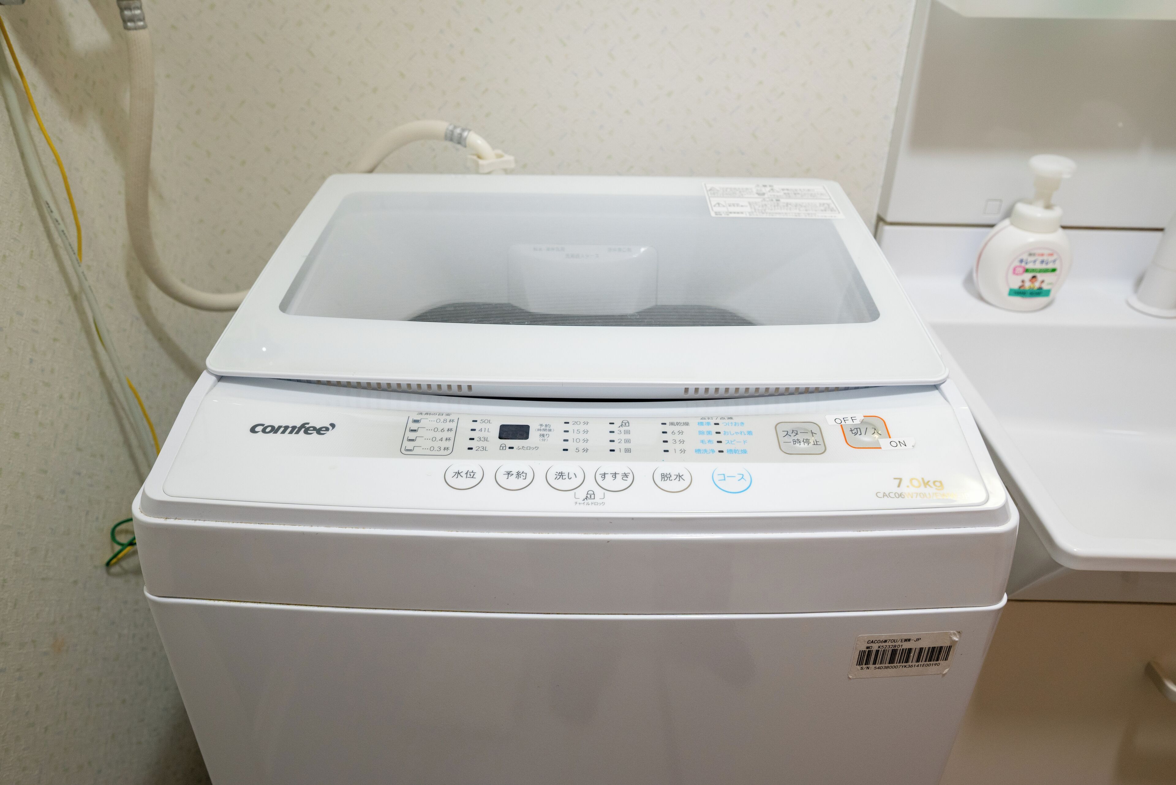 Een witte Comfee wasmachine met een bedieningspaneel, een wateraansluitslang en een fles handzeep op een witte aanrechtblad.