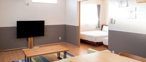 A compact hotel room with a flat-screen TV, a wooden dining table, a sofa, and a bed with a window view.