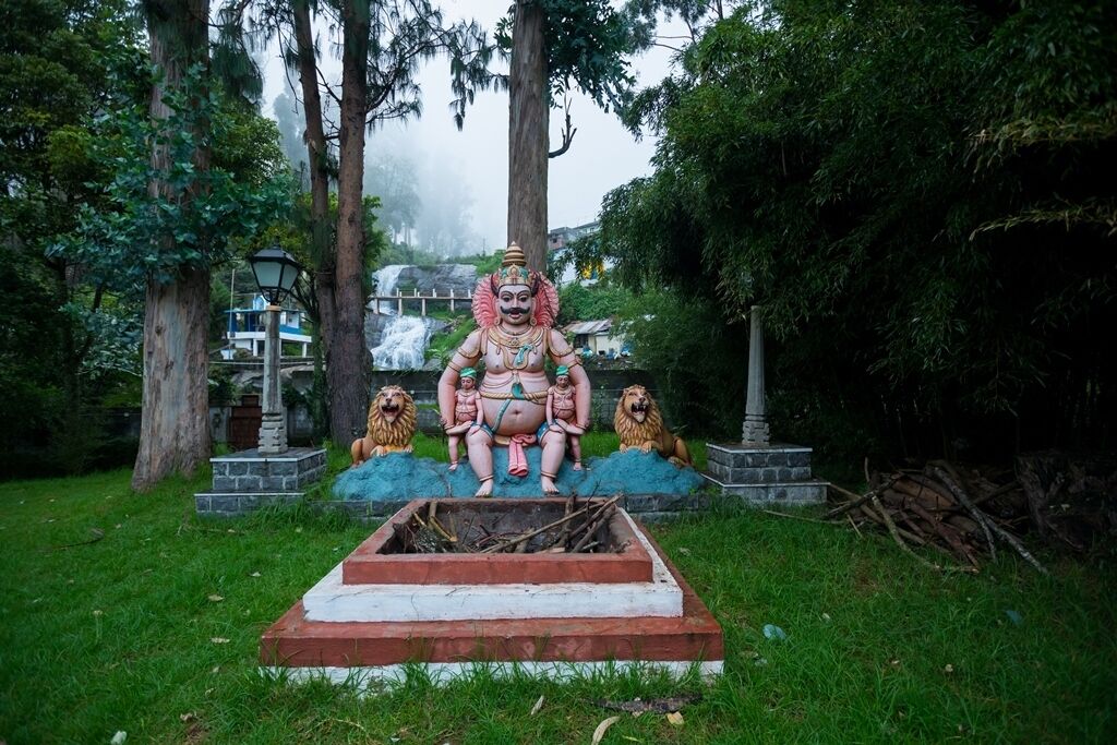 A statue of a deity with two lions, set on a stone platform in a park with trees and a waterfall in the background.