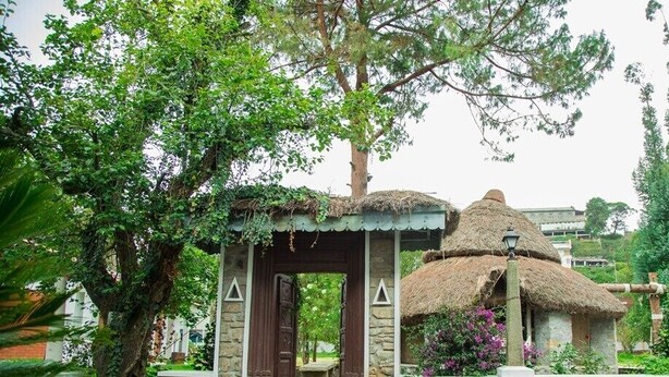 A traditional thatched-roof structure with a stone base, surrounded by lush greenery and trees. - Hotel Kodai International (Kodaikanal)
