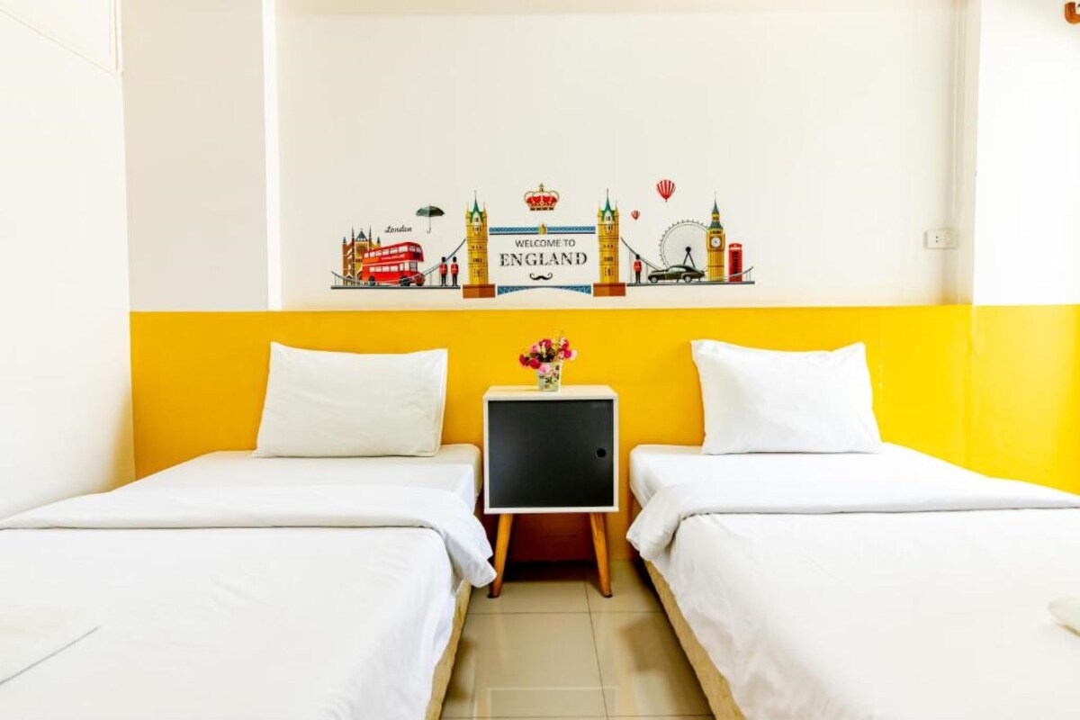 Ein Hotelzimmer mit zwei Betten, einer gelben Akzentwand, einem Schild mit der Aufschrift 'Welcome to England' und einem kleinen Tisch mit einem Blumenstrauß.