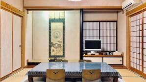 Um quarto japonês tradicional com tatame, uma mesa baixa com almofadas, uma televisão e portas deslizantes.