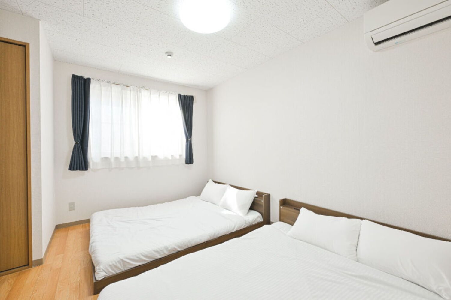 Une chambre avec deux lits simples, un plancher en bois, une fenêtre avec des rideaux et un appareil de climatisation.
