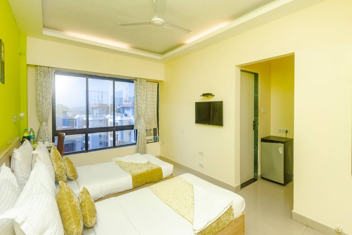 A hotel room with two beds, a ceiling fan, a window with curtains, a television, and a minibar.