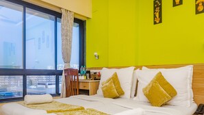 A hotel room with a large bed, a wooden nightstand, a desk with a chair, and a view of the city through a large window. - Tuliip Residency (Mumbai)