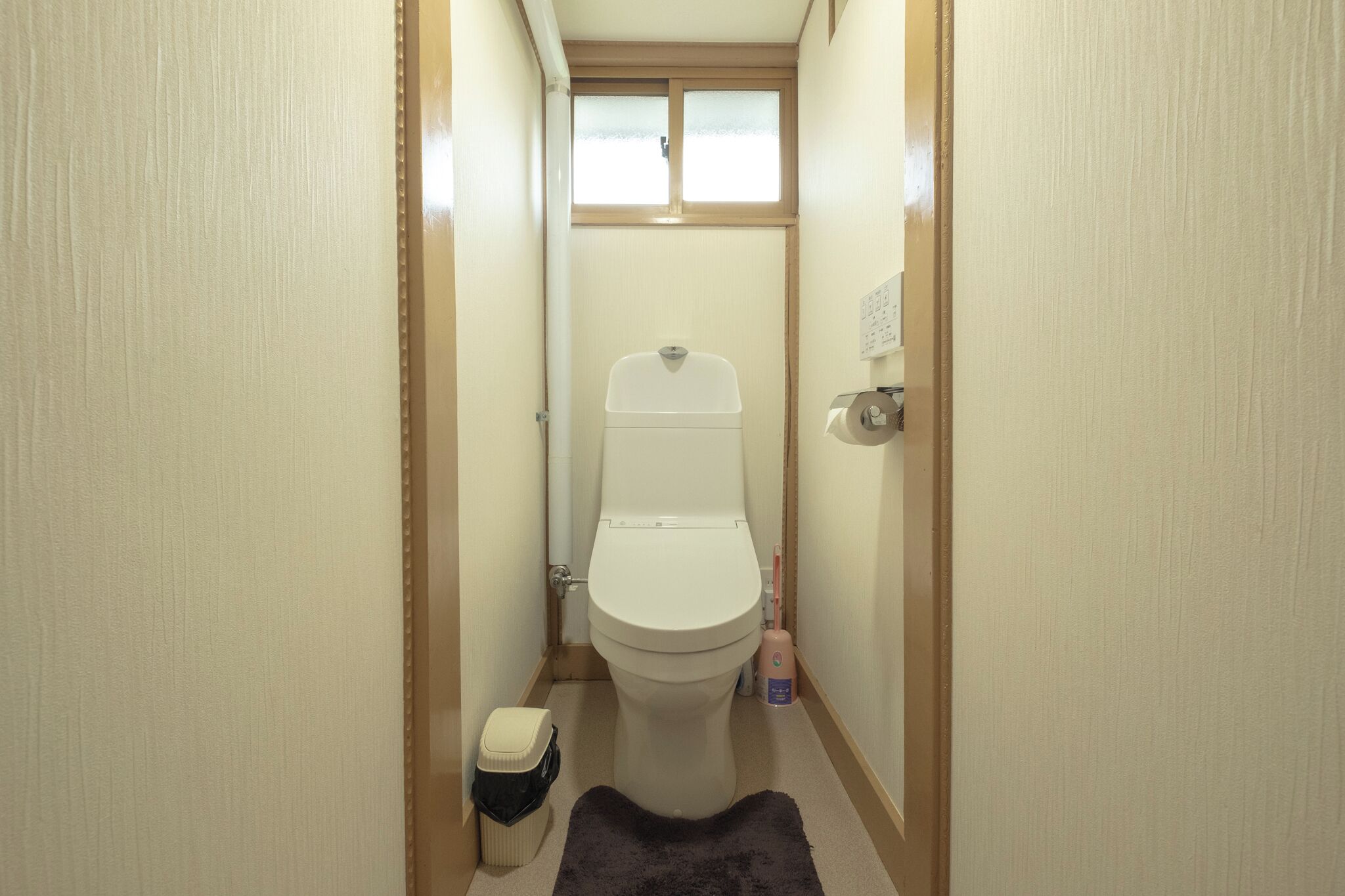 A narrow bathroom with a toilet, a small trash bin, and a bottle of hand soap on the floor.