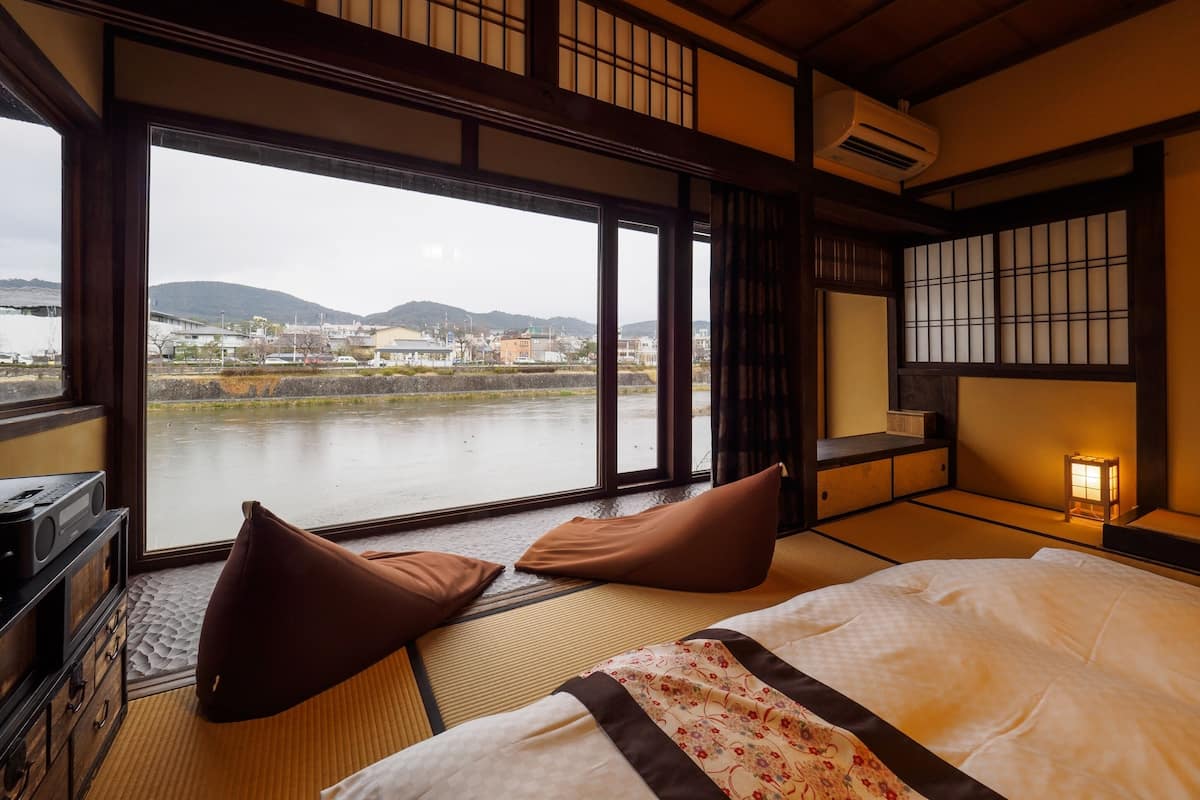 Una habitación tradicional japonesa con suelo de tatami, una cama con manta de estampado floral, un escritorio con radio y vistas a un río y edificios al fondo.