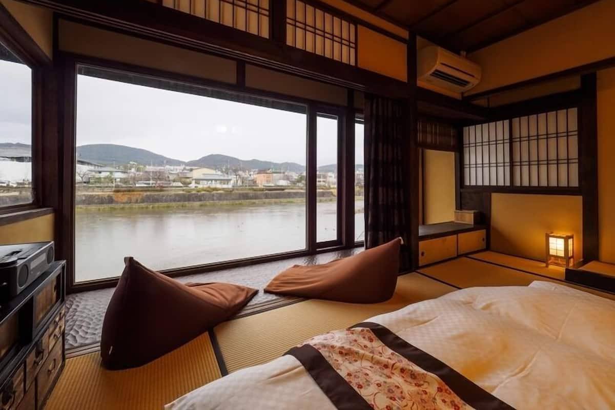 Una habitación tradicional japonesa con suelo de tatami, una cama con edredón floral, un escritorio con radio y vistas a un río y edificios al fondo.