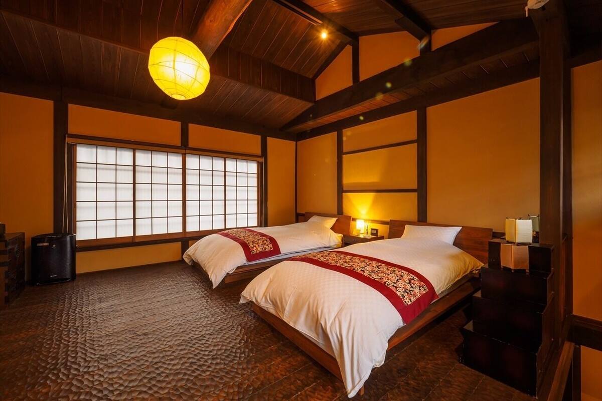 A traditional Japanese-style room with two beds, wooden beams, tatami flooring, and a large window.
