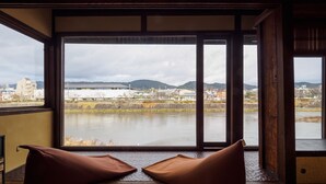A room with tatami flooring, a large window with a view of a river and distant buildings, and two brown cushions.