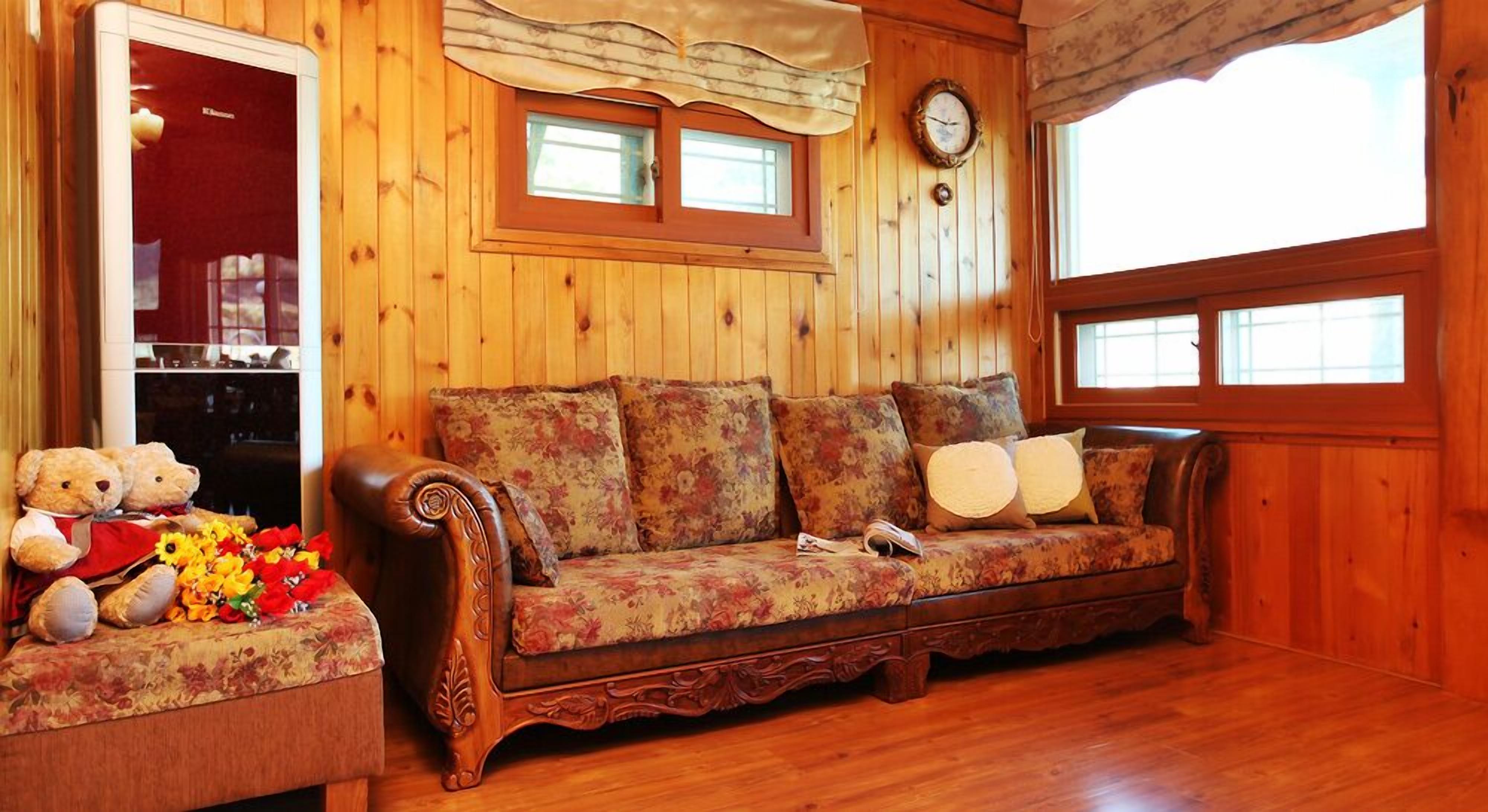 Un intérieur en bois avec un canapé, une petite table décorée d’ours en peluche et de fleurs, et une horloge au mur.