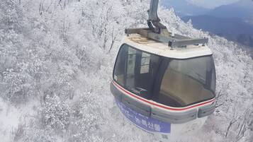 A cable car with a cabin and operator cabin, moving over a snow-covered landscape with trees and distant mountains.