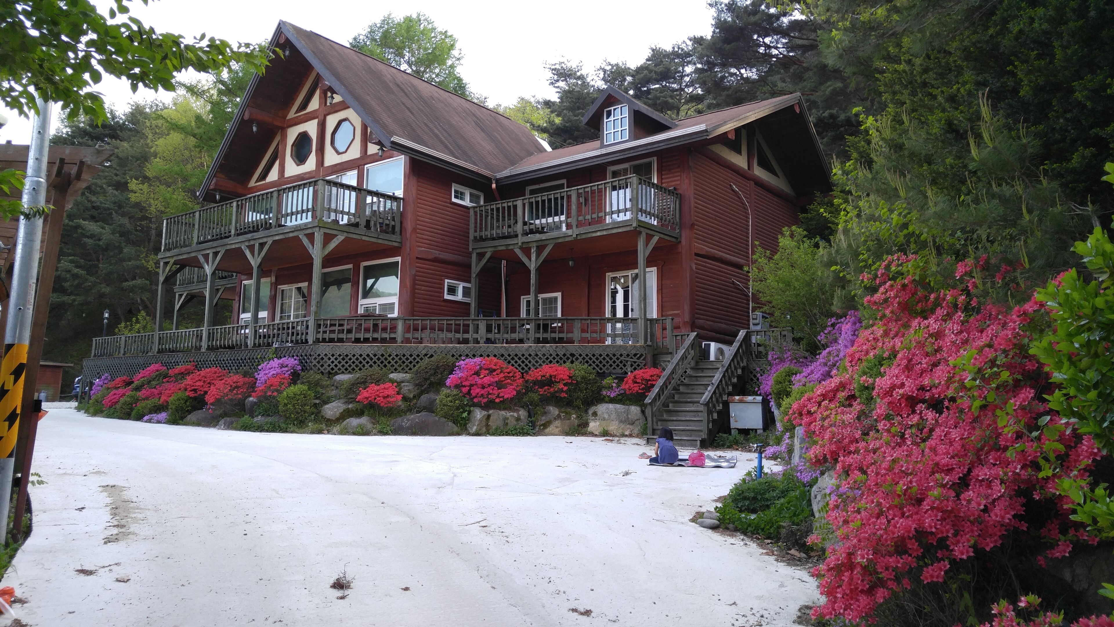 A large, two-story wooden house with a balcony, surrounded by lush greenery and flowering plants.