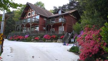A large, two-story wooden house with a balcony, surrounded by lush greenery and flowering plants.
