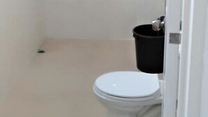Une petite salle de bain aux murs blancs, équipée de toilettes et d’une poubelle noire.