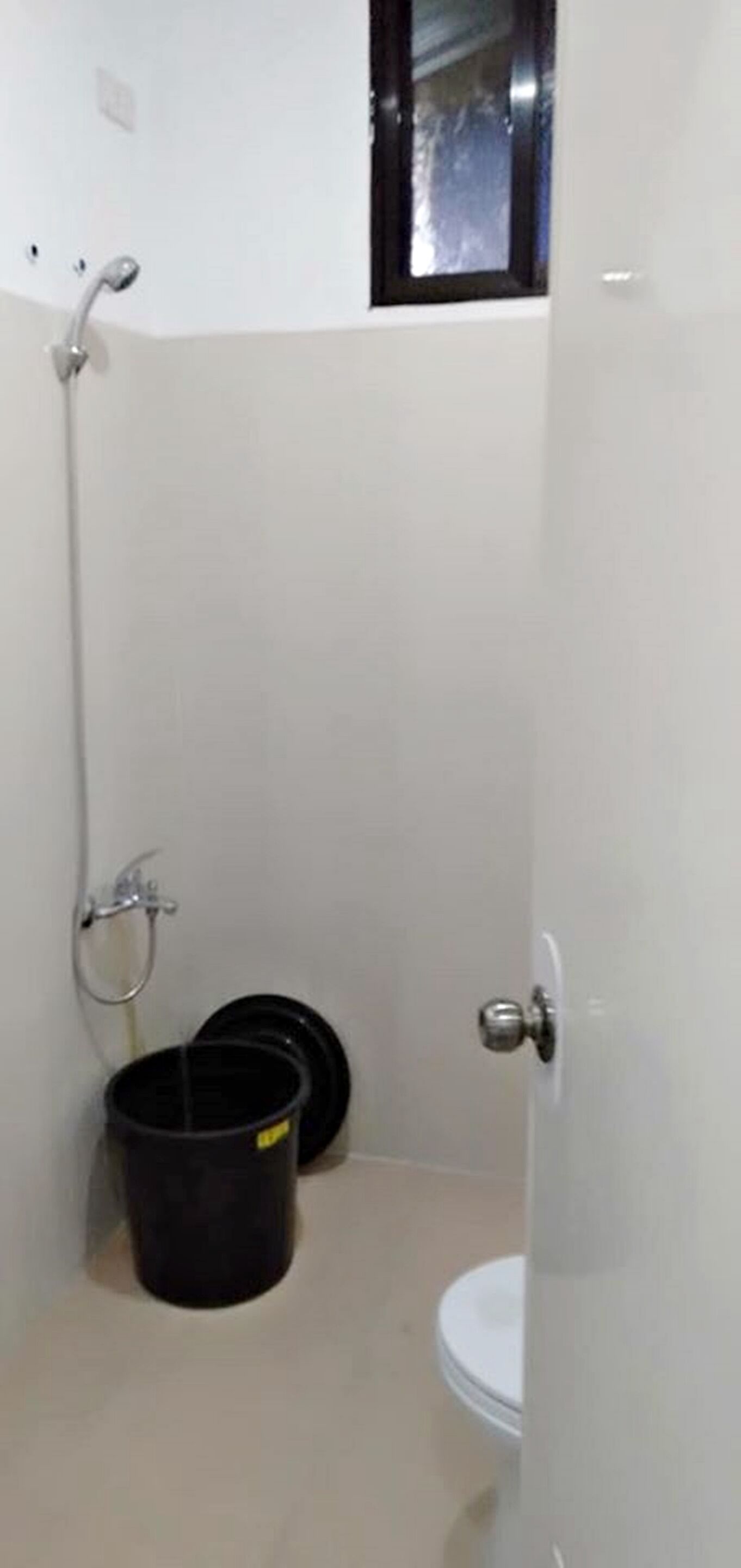 Un baño pequeño y cerrado con inodoro, zona de ducha y un cubo negro.