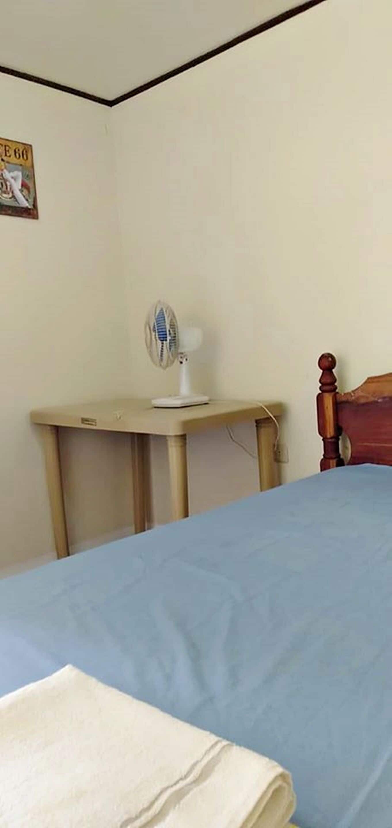A bedroom with a bed, a bedside table with a fan, and a wooden headboard.