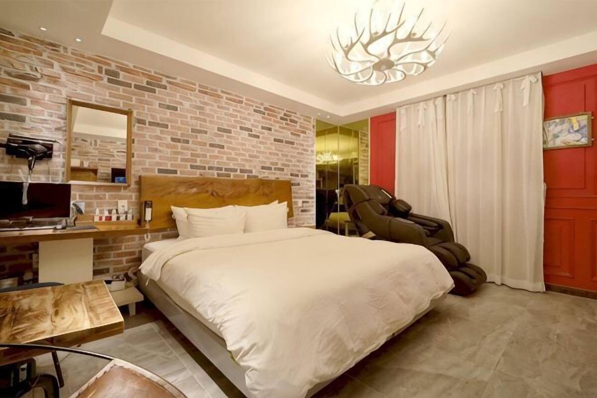 A hotel room with a brick wall, a bed, a desk, a chair, and a large window.