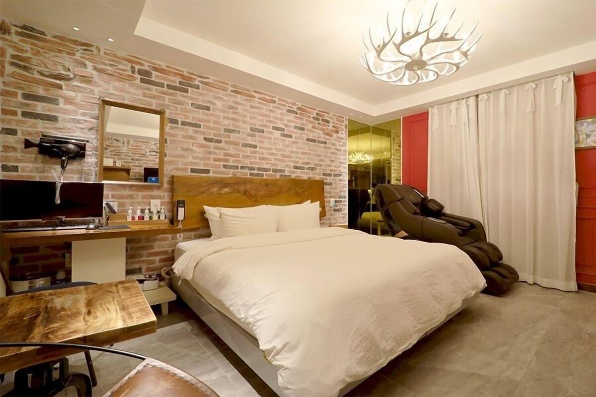 A bedroom with a brick wall, a bed, a massage chair, a TV, and a red door.
