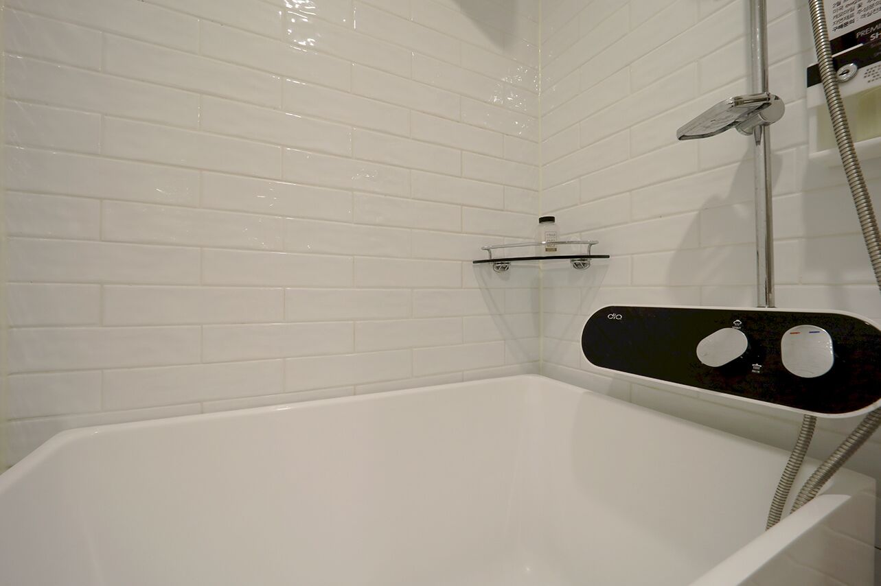 Un baño con azulejos blancos, una bañera, estante de vidrio y panel de control de ducha.