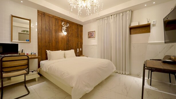 A hotel room with a large bed, wooden paneling, a chandelier, a desk, a chair, a TV, and a bathroom with a mirror and toilet. - February Hotel Busan Gangseo Annex Building  (Busan)