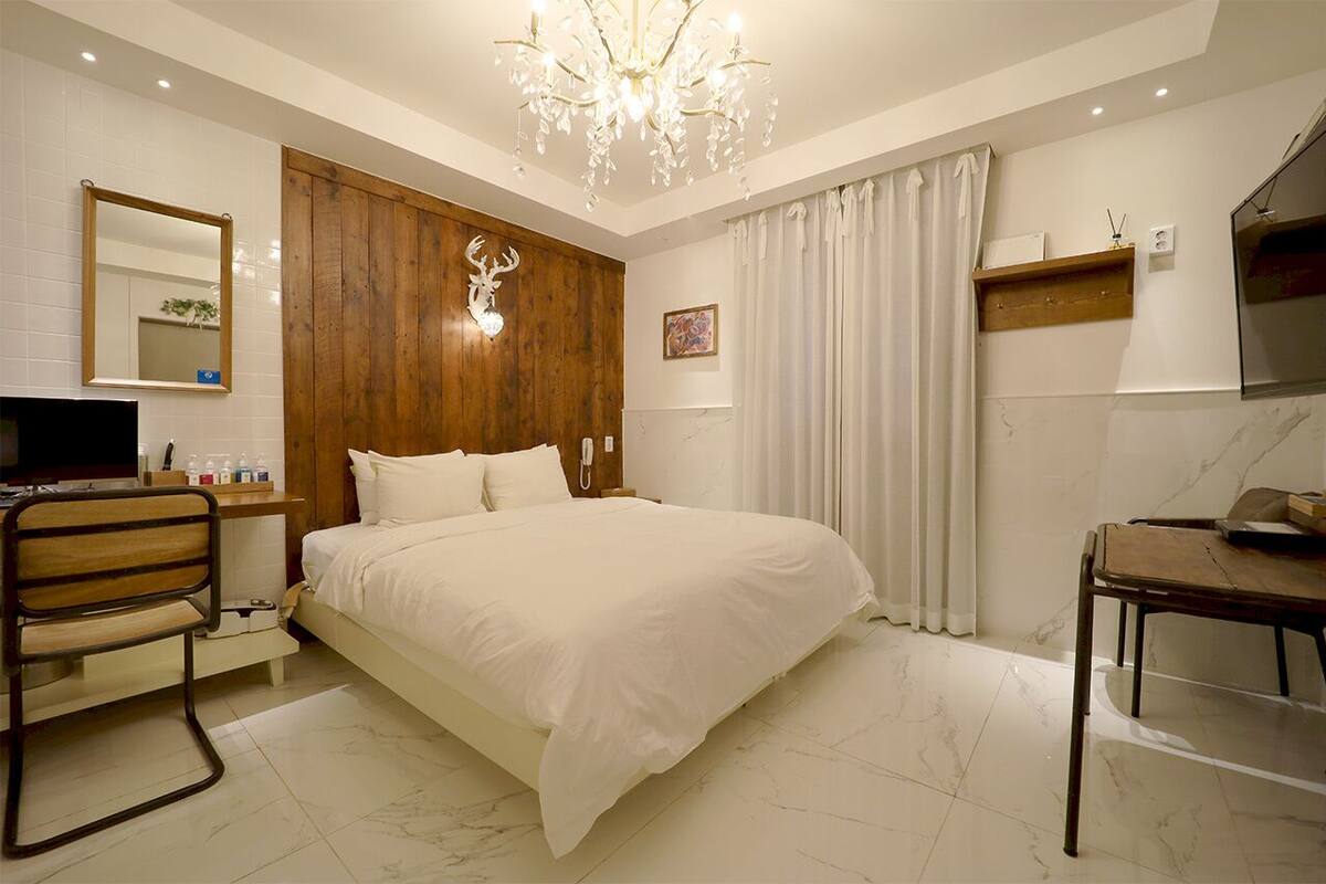 A hotel room with a large bed, wooden paneling, a chandelier, a desk, a chair, a TV, and a bathroom with a mirror and toilet.