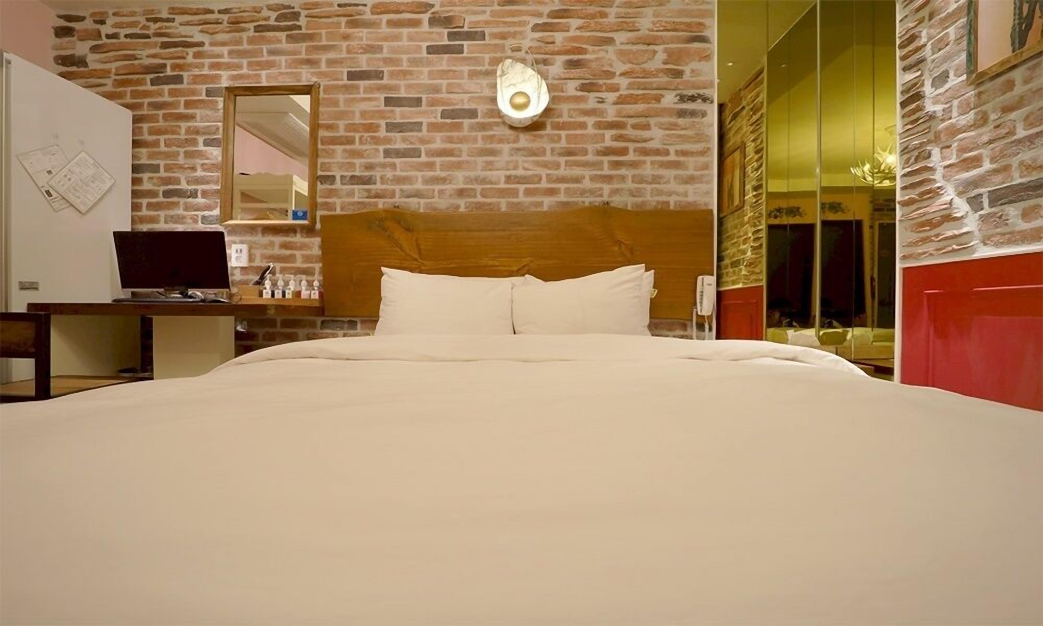 Habitación de hotel con pared de ladrillo, cabecero de madera, cama con ropa blanca, escritorio con computadora y espejo.