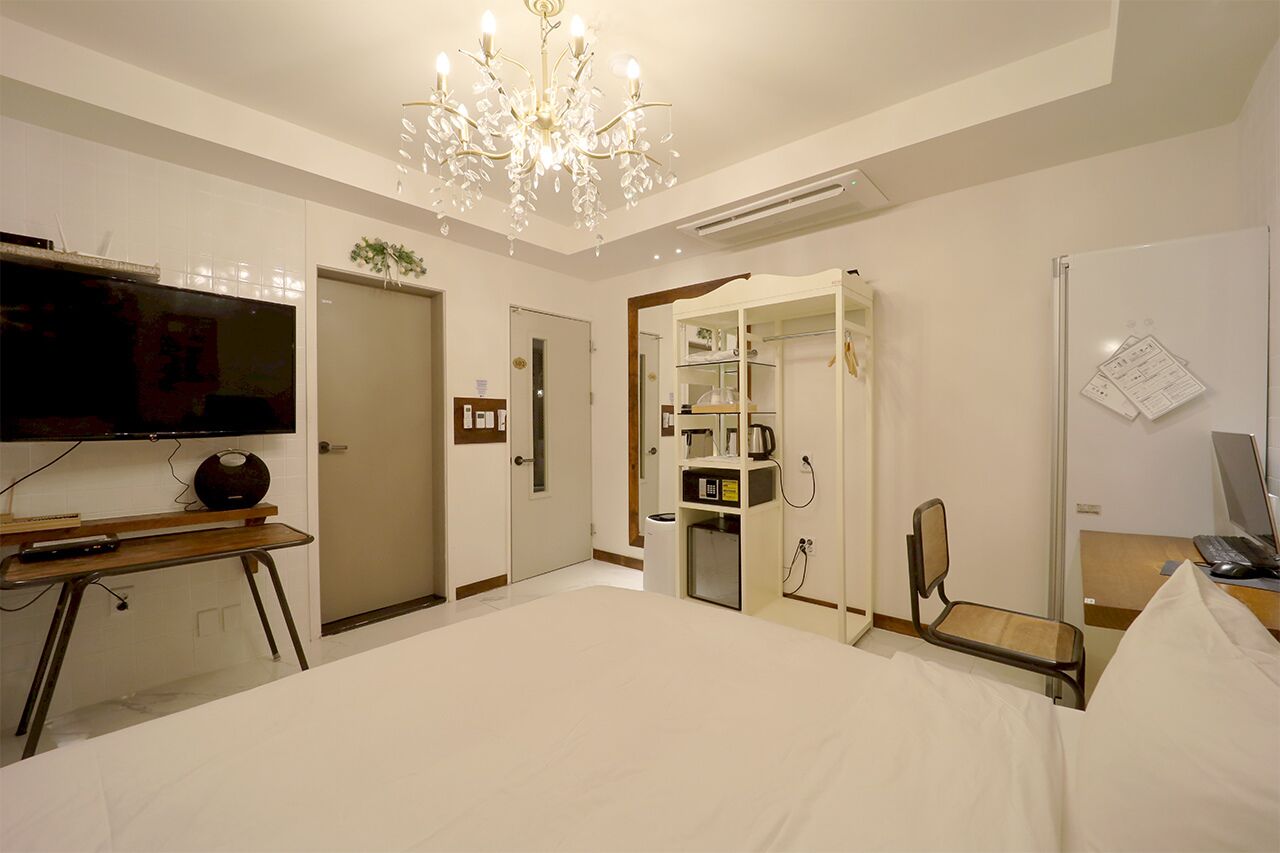 A hotel room with a large bed, a television, a desk with a computer, a chair, a mirror, and a chandelier.