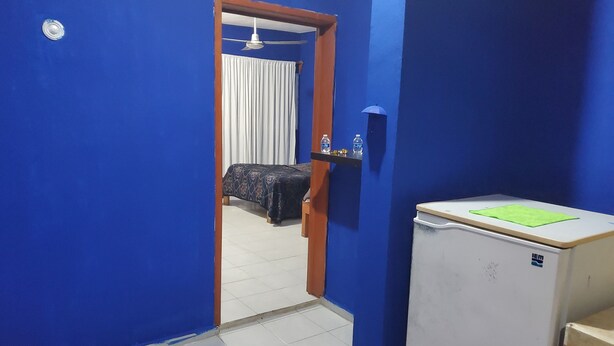 Een kamer met blauwe wanden, een bed met een bedspreider met patroon, een klein tafeltje met een groene onderzetter en een witte koelkast.