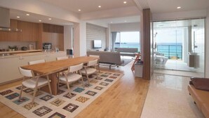 A modern kitchen with wooden cabinets, a dining area with a wooden table and white chairs, and a large sliding glass door leading to a balcony with a view of the sea.