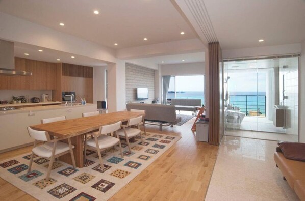 A modern kitchen with wooden cabinets, a dining area with a wooden table and white chairs, and a large sliding glass door leading to a balcony with a view of the sea. - K Seaveiw 250㎡ ONNA 2 big suites, 3 full bath 3F (Nakadomari, Onna, Kunigami-gun)