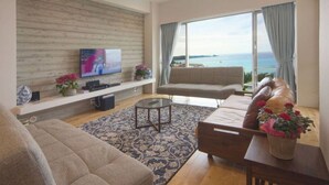 A modern living room with a large TV, comfortable seating, a wooden wall, and a view of the sea.