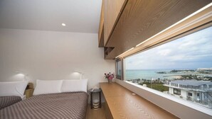 A modern hotel room with two beds, a wooden bench, a small table, and a large window offering a view of the sea and buildings.