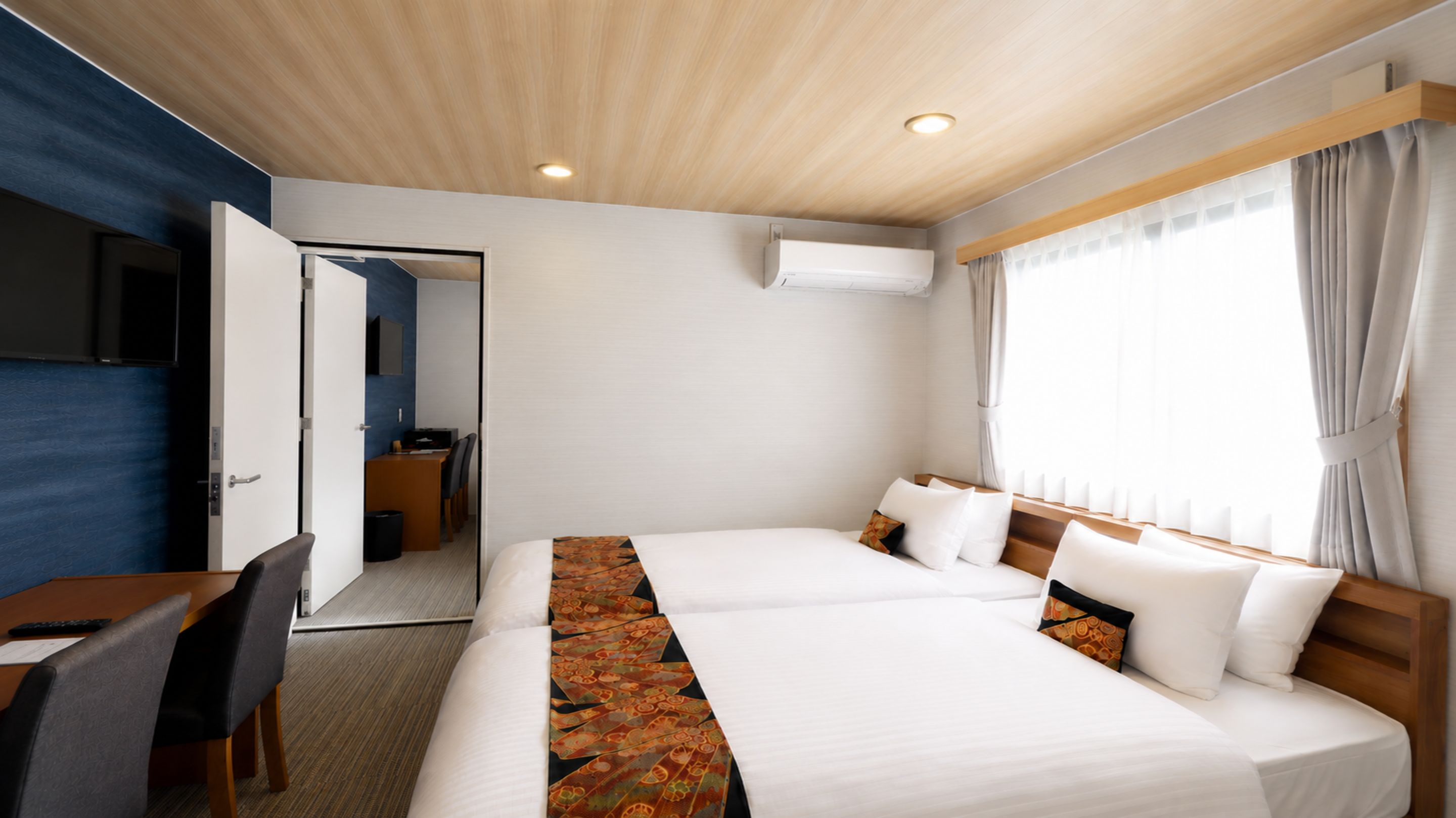 Quarto de hotel com cama, escrivaninha, cadeira e televisão.