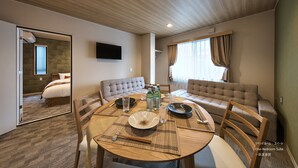 Suite de una habitación con comedor, sofá, televisor montado en la pared y ventana con cortinas.
