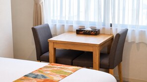 Een hotelkamer met een houten tafel, twee stoelen, een bed met een bloemenprint en een raam met dunne gordijnen.