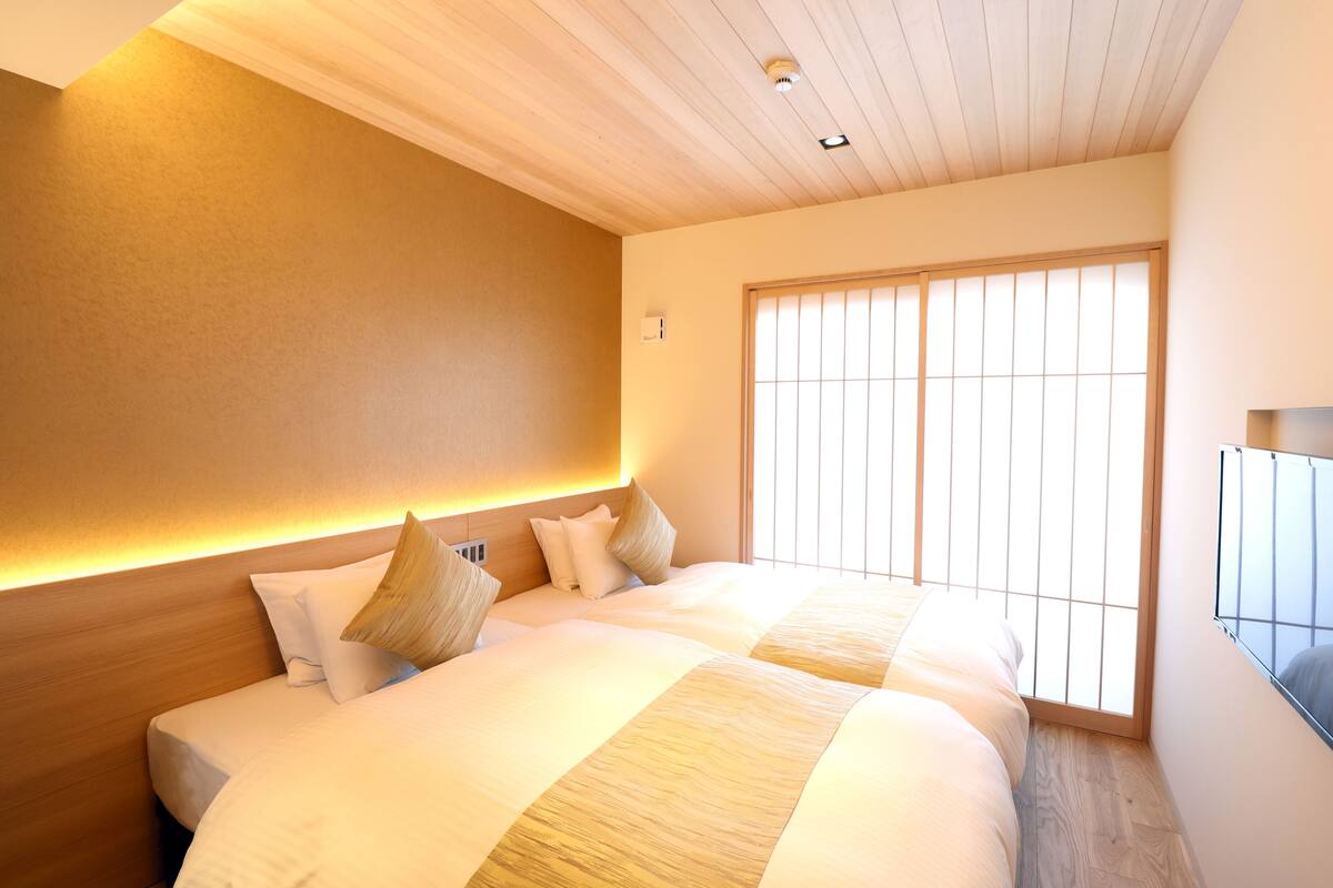 A hotel room with a large bed, a wooden headboard, a large window with blinds, and a flat-screen TV mounted on the wall.