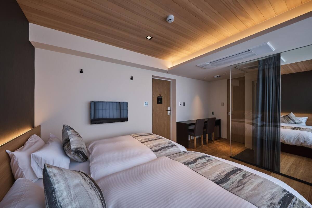A modern hotel room with a large bed, a desk, a television, and a sliding door leading to a bathroom.