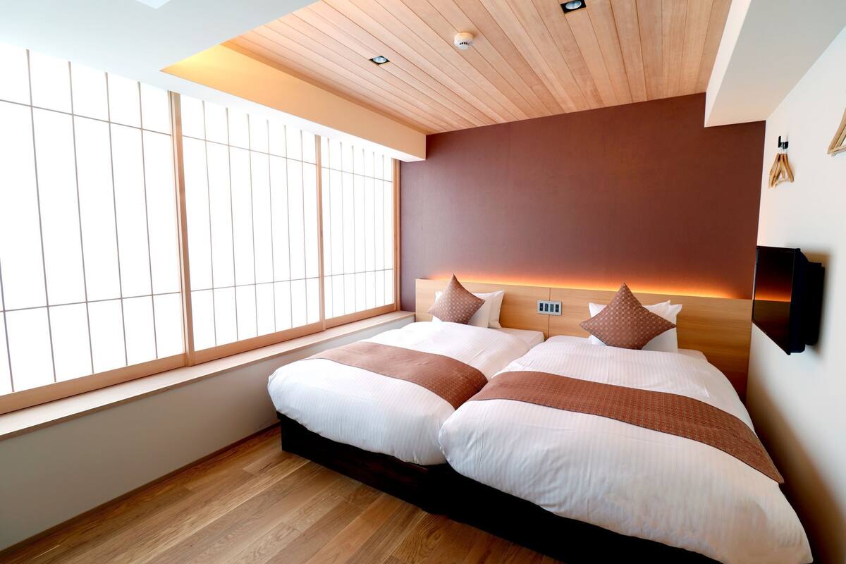 A modern hotel room with a large bed, wooden flooring, a large window with blinds, and a wooden ceiling.