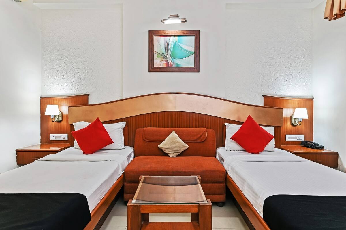 Habitación de hotel con dos camas individuales, cabecero de madera, lámparas de noche, una mesita pequeña y un cuadro en la pared.