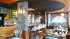 Interior de un restaurante con mesas y sillas de madera, una columna central con un envoltorio de periódico, y una pared colorida con nombres de ciudades.