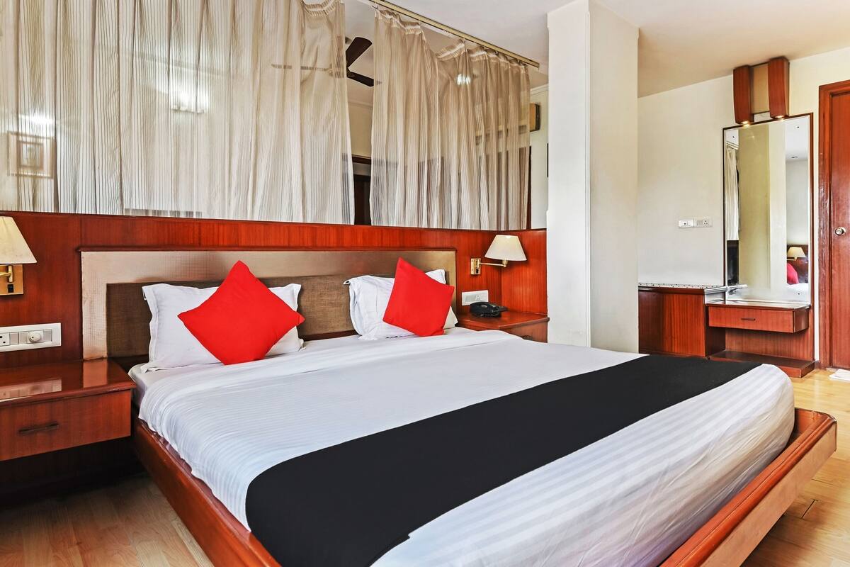 Habitación de hotel con una cama grande, cabecera de madera, dos mesitas de noche con lámparas, un espejo y un aparador de madera.