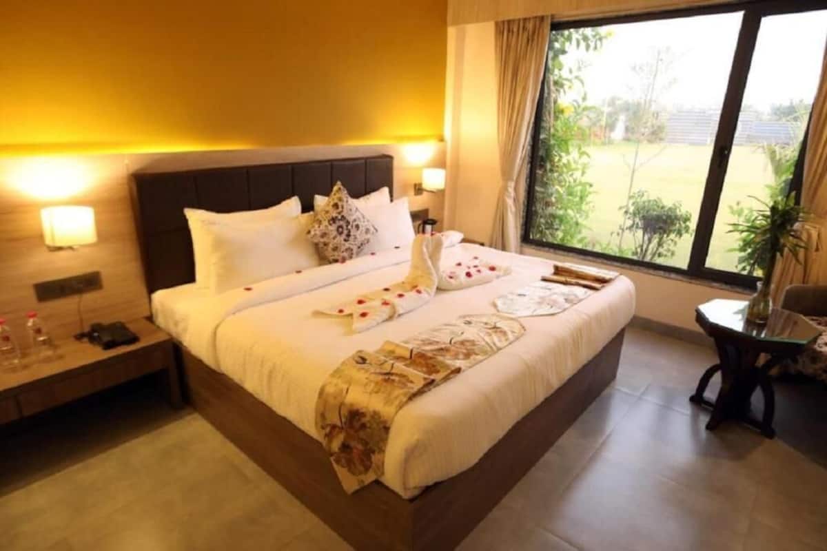 Habitación de hotel con una cama grande, mesitas de noche, una ventana con vistas a la vegetación y una pequeña mesa con una silla.