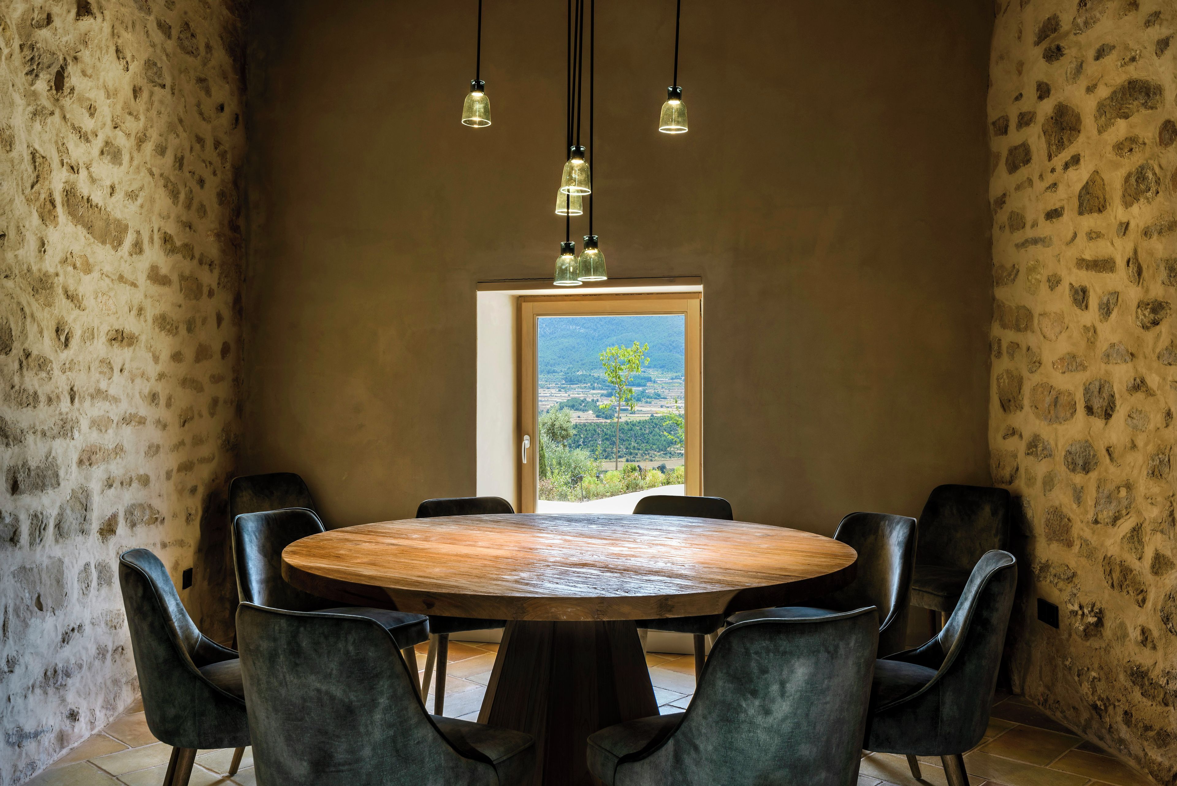 A round wooden table with six chairs, a stone wall, and a view through a doorway.