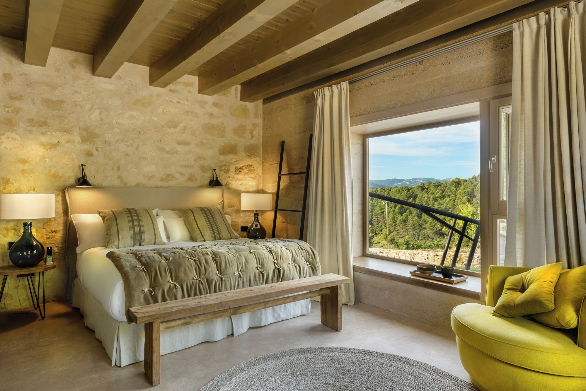 Un dormitorio con pared de piedra, una cama con una banca, un sillón amarillo y vista al exterior.