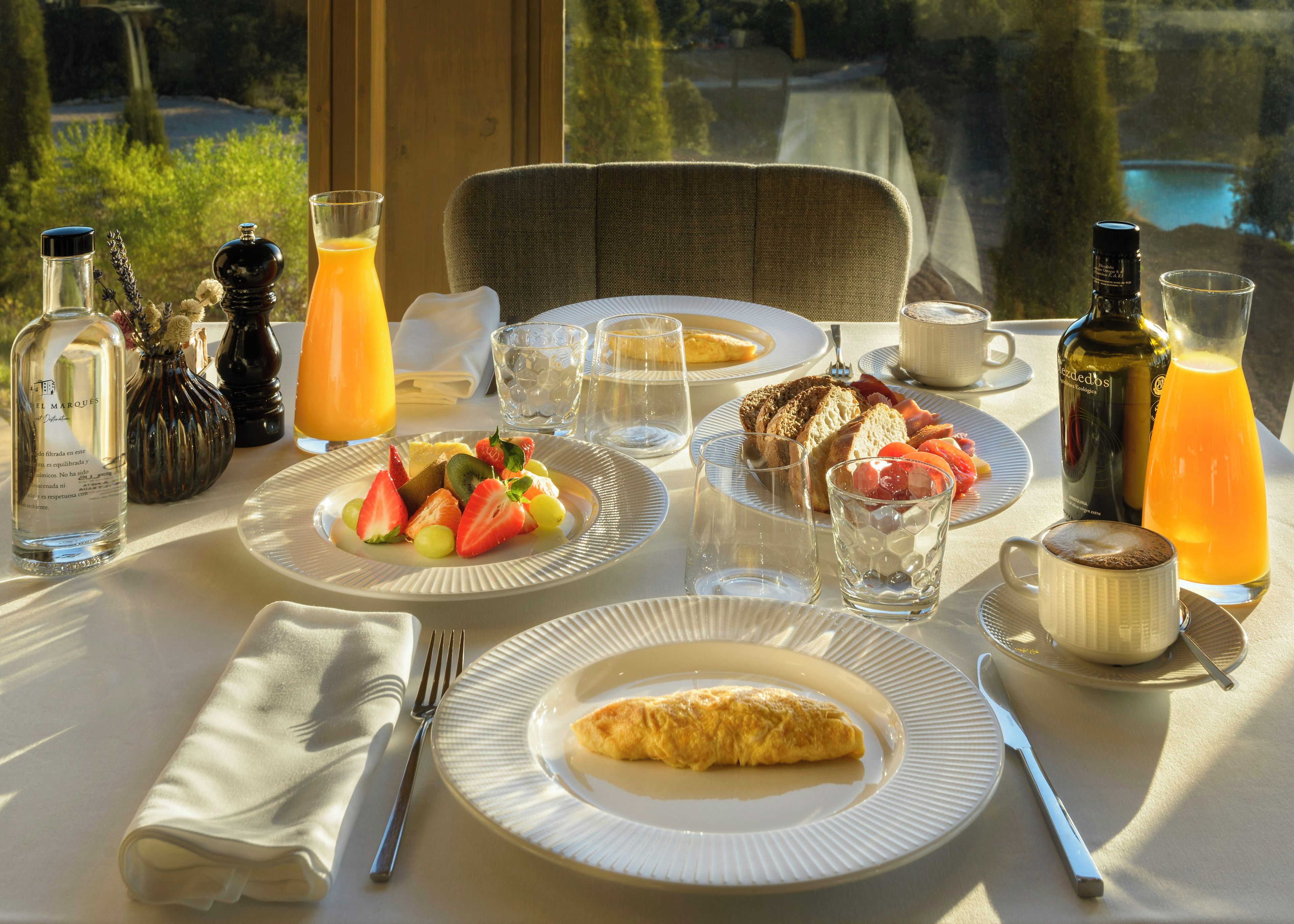 Desayuno con platos de comida, vasos y botellas sobre una mesa con vistas a vegetación y un cuerpo de agua.