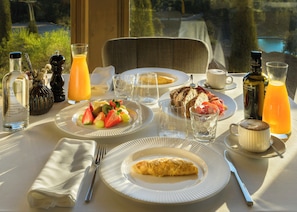 Una colazione servita con piatti di cibo, bicchieri e bottiglie su un tavolo affacciato su un paesaggio verde e un corso d'acqua.