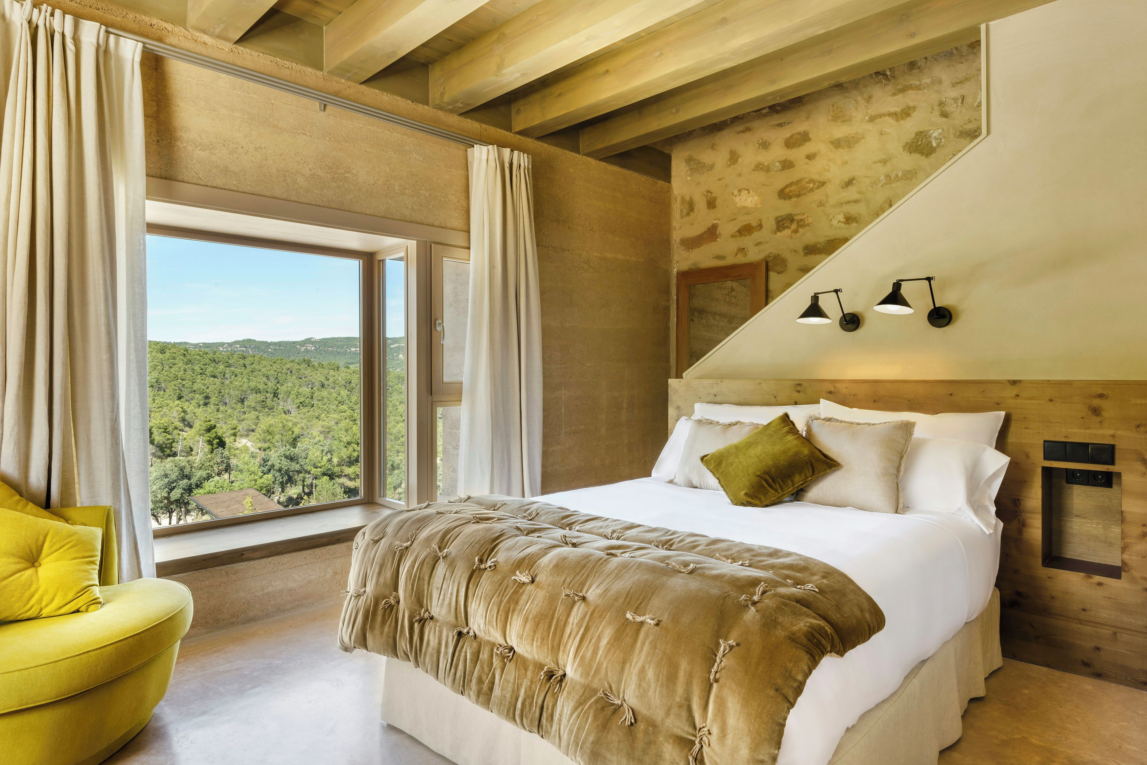 A bedroom with a large bed, a yellow armchair, and a view of a forest through a window.