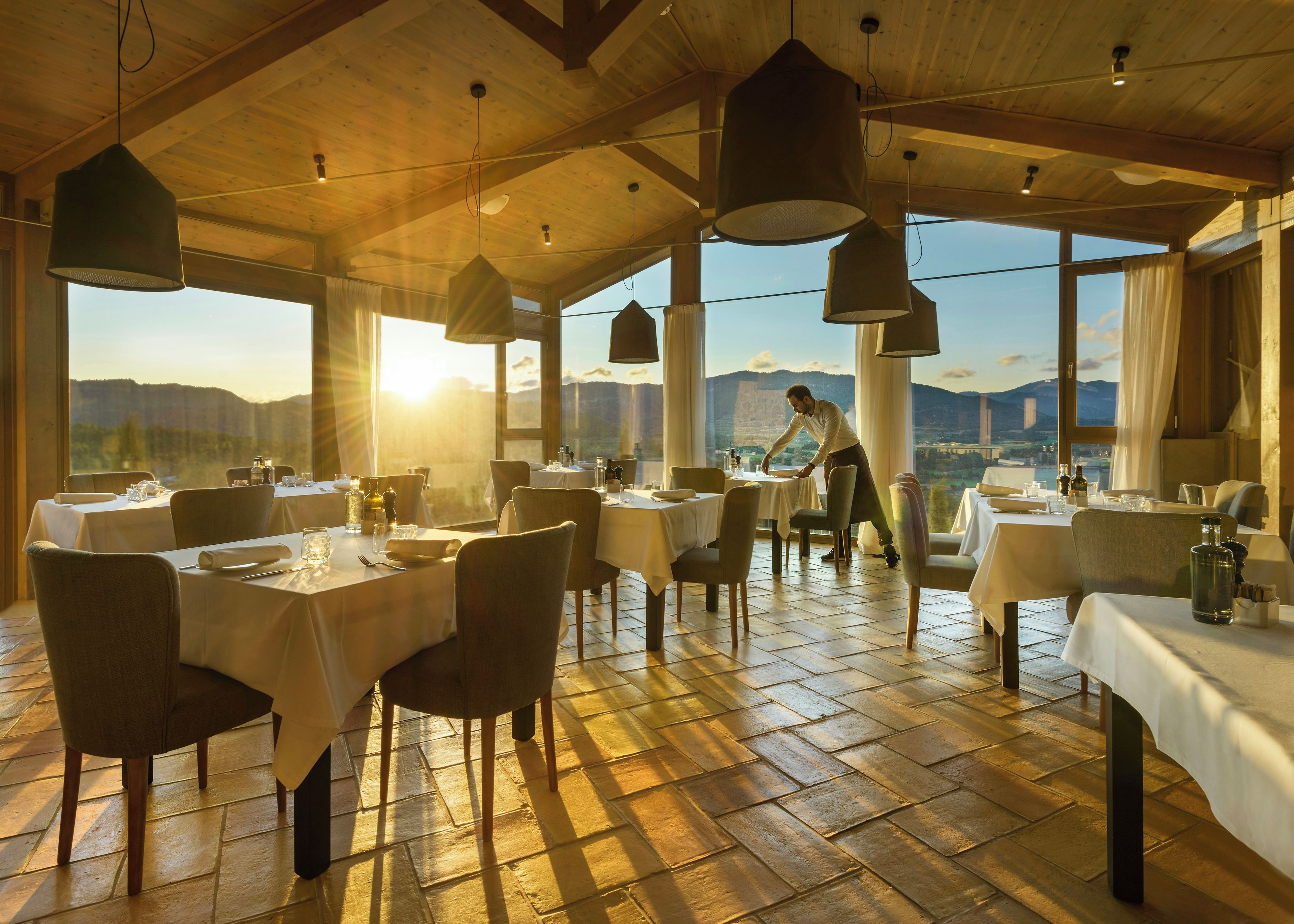 En restaurang med dukade bord, en servitör som tar hand om gästerna och en vacker utsikt över bergen genom stora fönster.